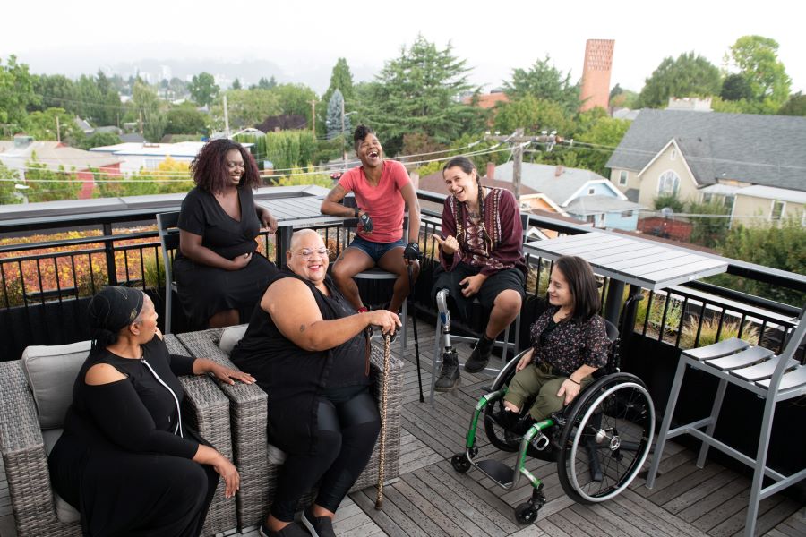 Overhead shot of six disabled people of color at a rooftop deck party. An Indigenous Two-Spirit person with a prosthetic leg smiles directly at the camera and gives a thumbs up while everyone else is engaged in conversation